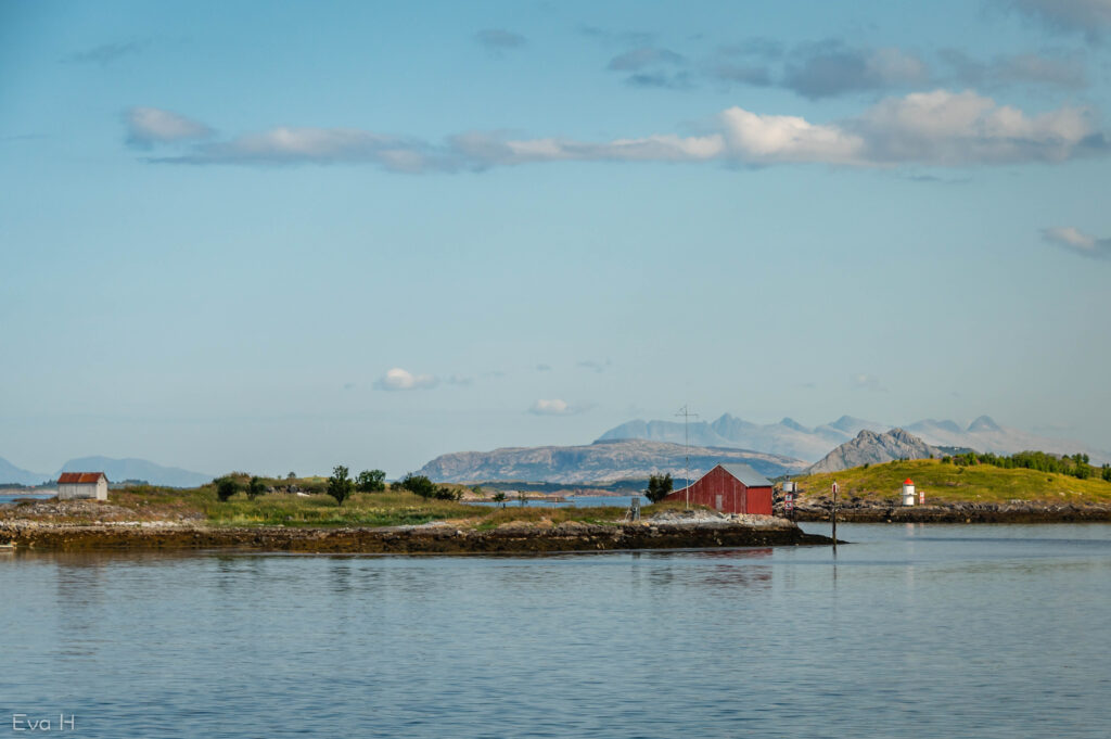 Naust og fyrlykt på holmen en vakker sommerdag, med de sju søstrene i bakgrunnen. Fargebilde. Fotografi.