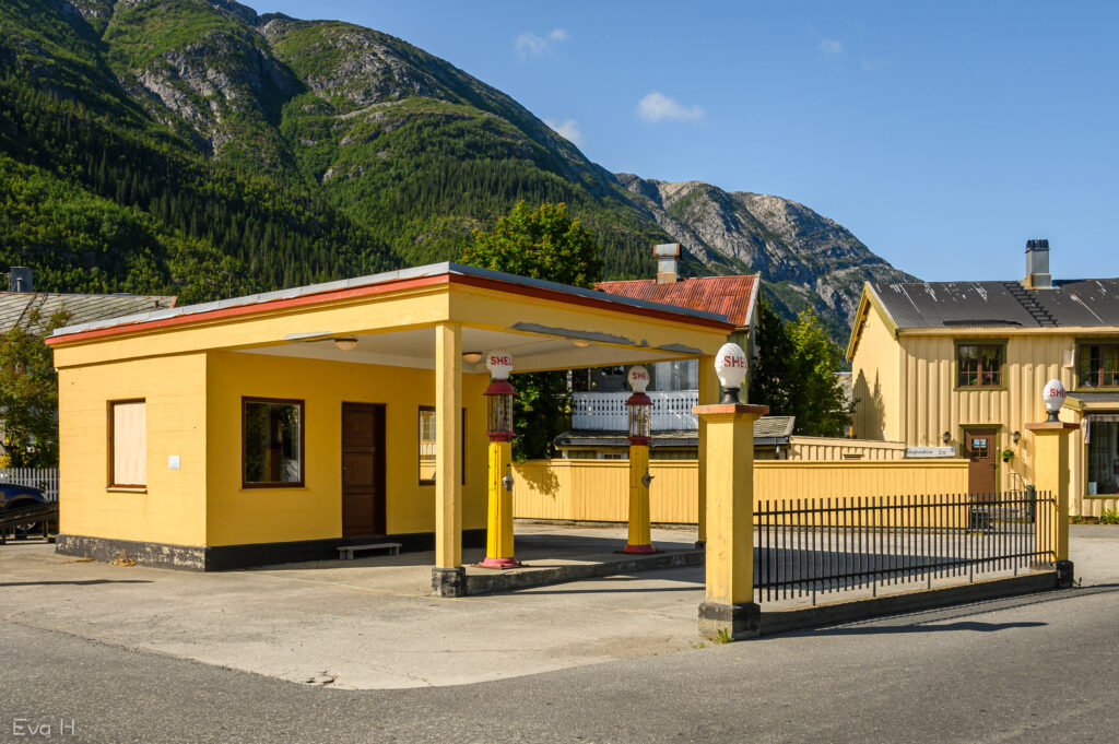 Nedlagt retro Shell-stasjon med fjell bak. Gule hus, blå himmel. Fargebilde. Fotografi.