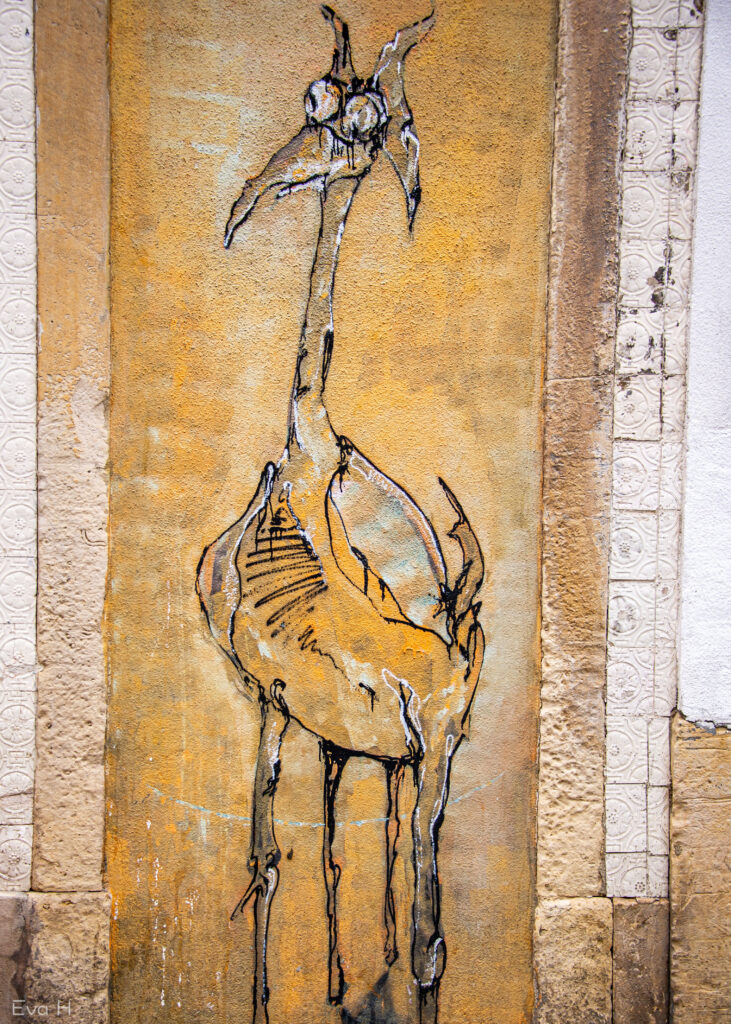 Sjiraff-liknende dyr med store øyne ser forundret på verden. Gule jordtoner, malt på mur. Gatekunst. Lisboa. Fotografi