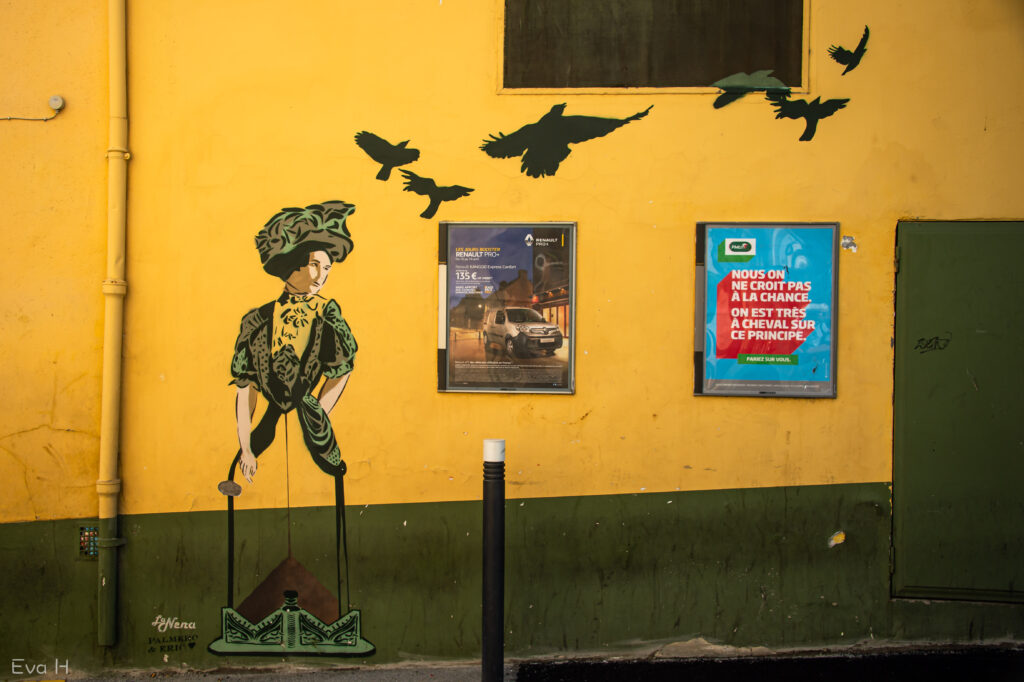 Gul og grønn dame med hatt og fugler på en vegg i Frankrike. Gatekunst