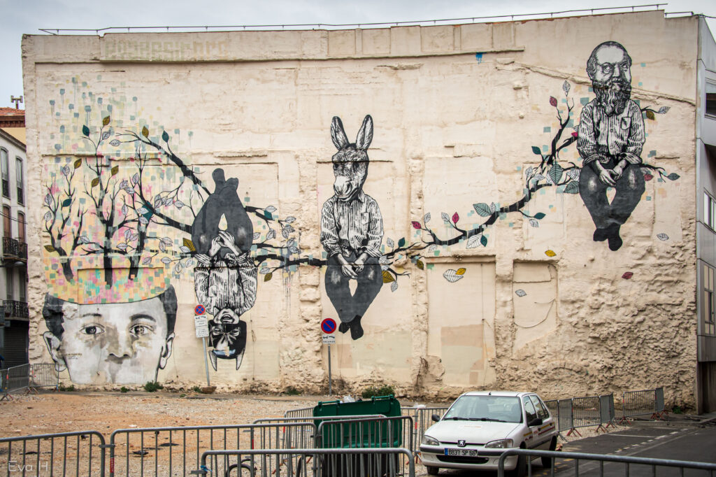 Stor murvegg over flere etasjer dekorert med blomstrende gren. Mann med skjegg,mann med eselhode sitter på grenen. MAnn med Batman-maske henger opp ned. Fotografi av gatekunst