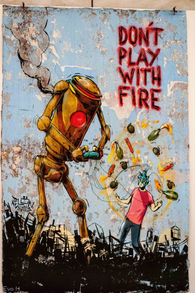 Robot styrer menneske med spillkontroll. Granater og dynamitt rundt mennesket. Tekst: Don't play with Fire. Fotografi av gatekunst
