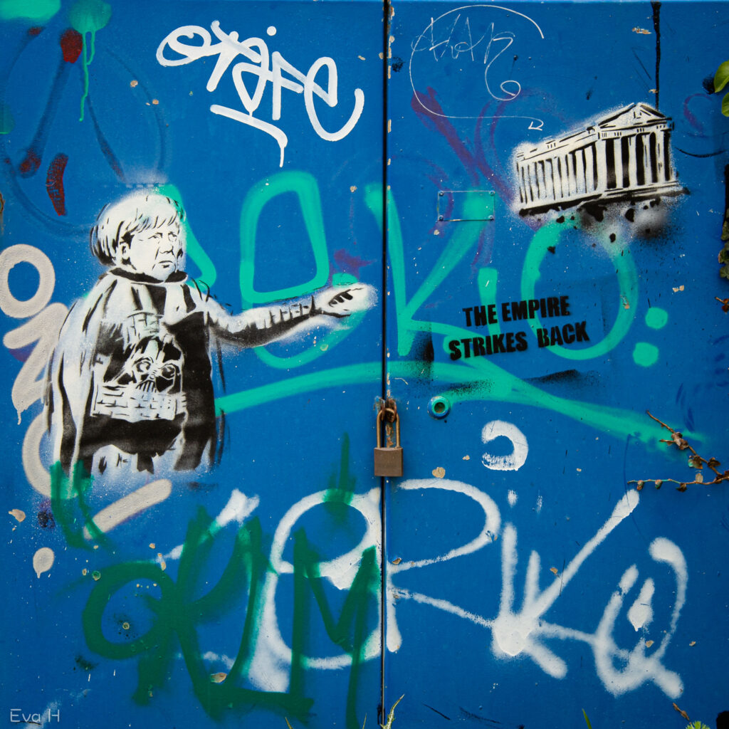A. Merkel kledd som figur i Star Wars på blå metalldør. Tekst: The Empire strikes back. Fotografi av gatekunst