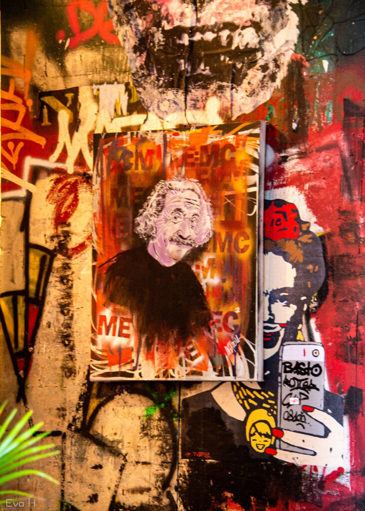 Vegg med mange detaljer, orange og rødt dominerer. Einstein-liknende person smiler under stygg kjeft. Fotografi av gatekunst